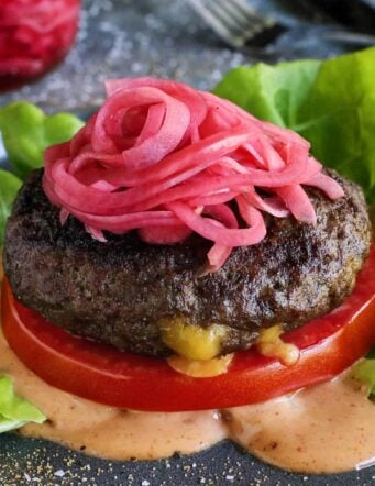 a Juicy Lucy cheese-stuffed burger on a plate, with lettuce, pickled red onions, slice of tomato, and creamy burger sauce