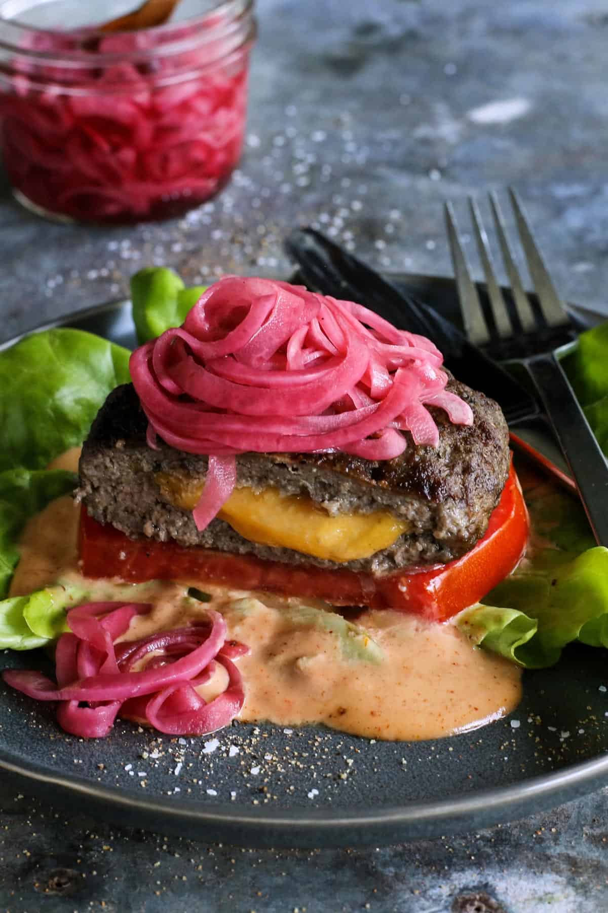 A Juicy Lucy burger is a juicy patty stuffed with molten, melty cheese. Served on a low-carb lettuce wrap or bun with your favorite toppings! a Juicy Lucy cheese-stuffed burger on a plate, with lettuce, pickled red onions, slice of tomato, and creamy burger sauce