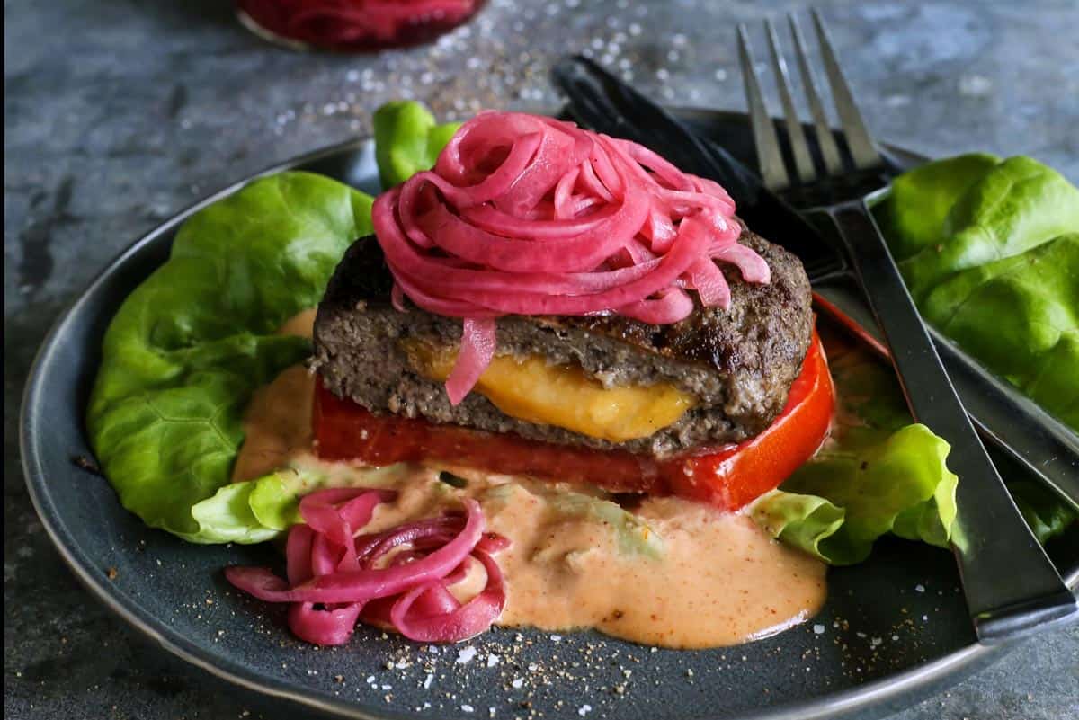 A Juicy Lucy burger is a juicy patty stuffed with molten, melty cheese. Served on a low-carb lettuce wrap or bun with your favorite toppings! a Juicy Lucy cheese-stuffed burger on a plate, with lettuce, pickled red onions, slice of tomato, and creamy burger sauce