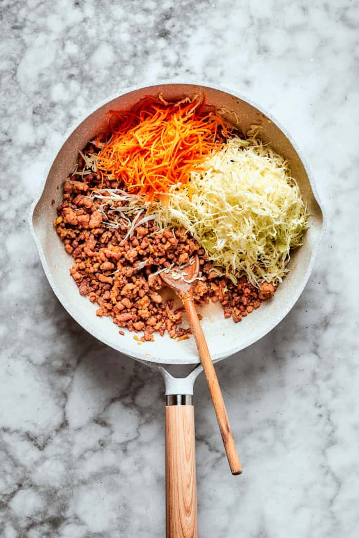 This easy Egg Roll in a Bowl recipe is loaded with ground pork, cabbage and carrots. Served with crispy wonton strips and a tasty Asian sauce! Overhead view of cabbage and carrots added to ground pork in skillet
