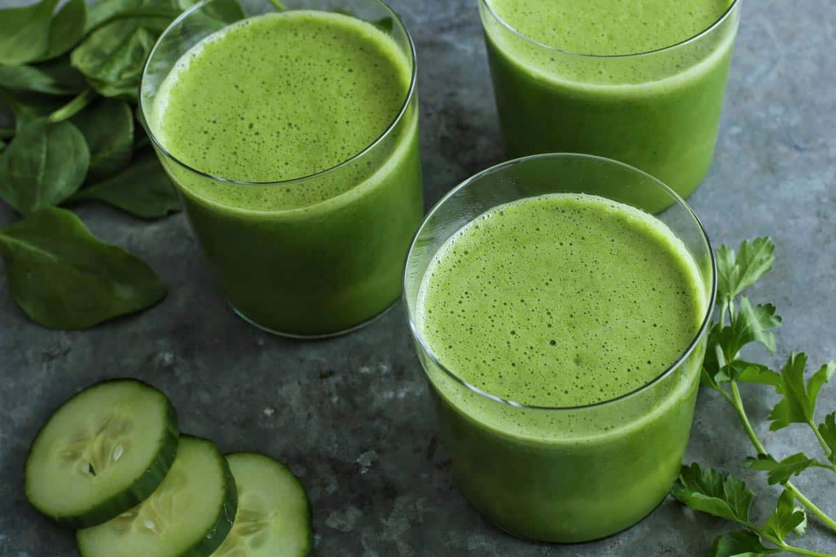 Green Juice is an easy blender recipe that whirls together spinach, parsley, cucumber, celery, green bell pepper, and orange juice. three glasses of green juice with spinach, cucumber slices, and parsley on the side