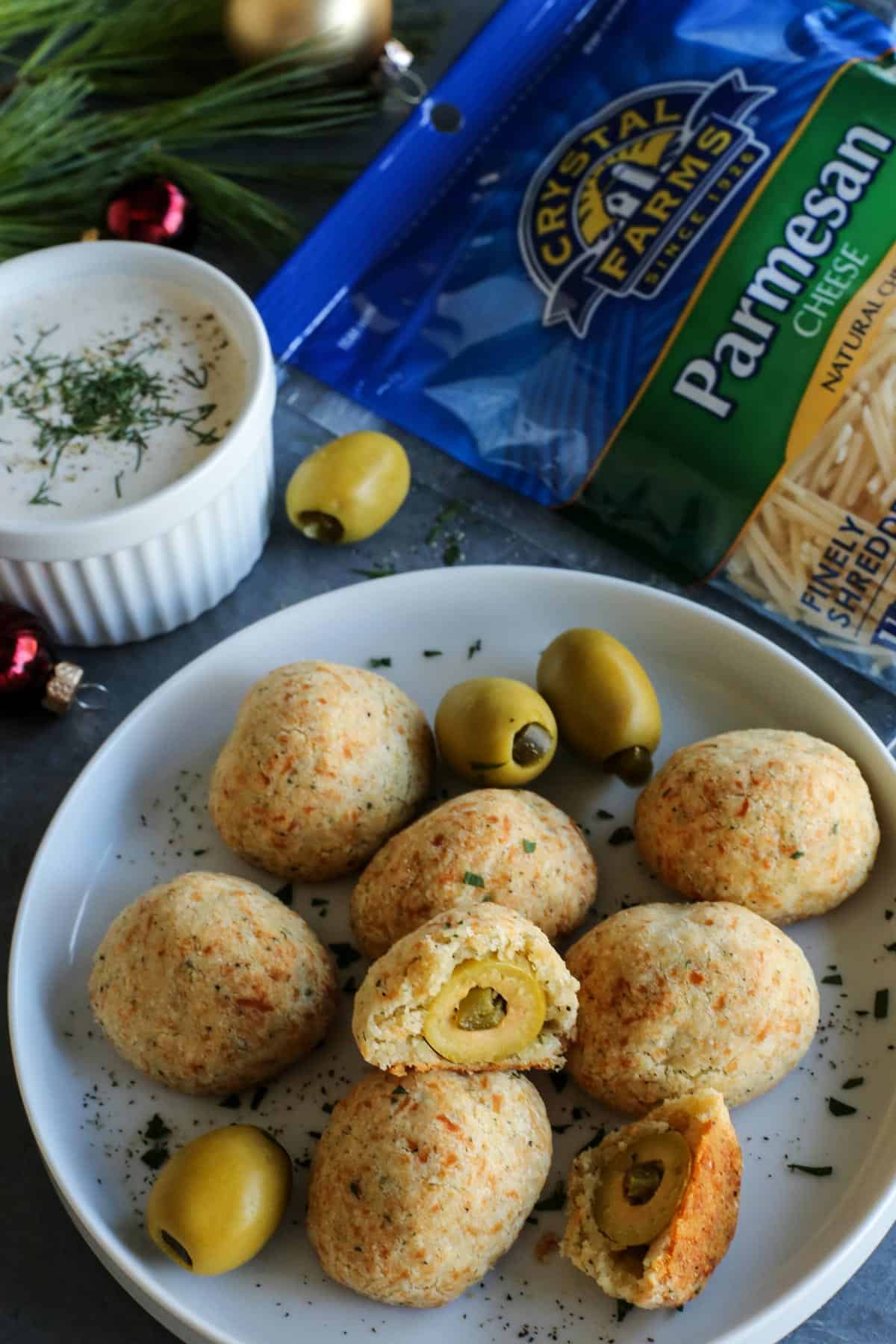 Olive Cheese Balls feature flaky, tender, cheesy dough that surrounds a warm and briny olive for a fun and easy appetizer. baked olive jalapeno cheese balls on a plate - with a package of Crystal Farms Parmesan cheese and a cup of buttermilk ranch dip on the side