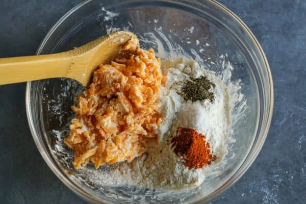 Olive Cheese Balls feature flaky, tender, cheesy dough that surrounds a warm and briny olive for a fun and easy appetizer. shredded sharp cheddar cheese, butter, flour, paprika, salt, and black pepper in a clear bowl with a wooden spoon