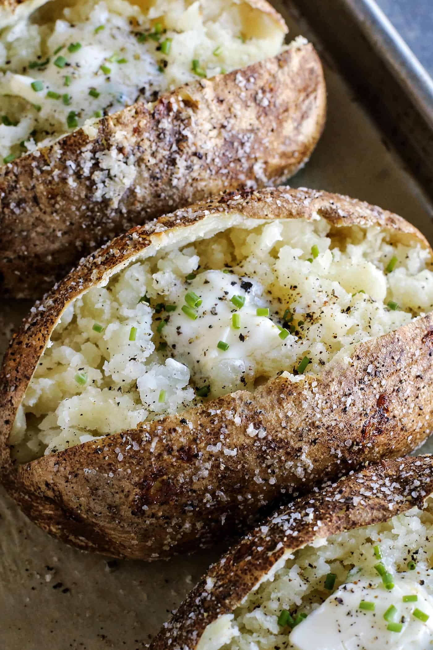 This baked potato recipe makes fluffy, tender potatoes with salty, crispy skin! Learn how to bake potatoes in the oven, plus 3 more ways! baked potatoes on pan, split open, with butter and chives