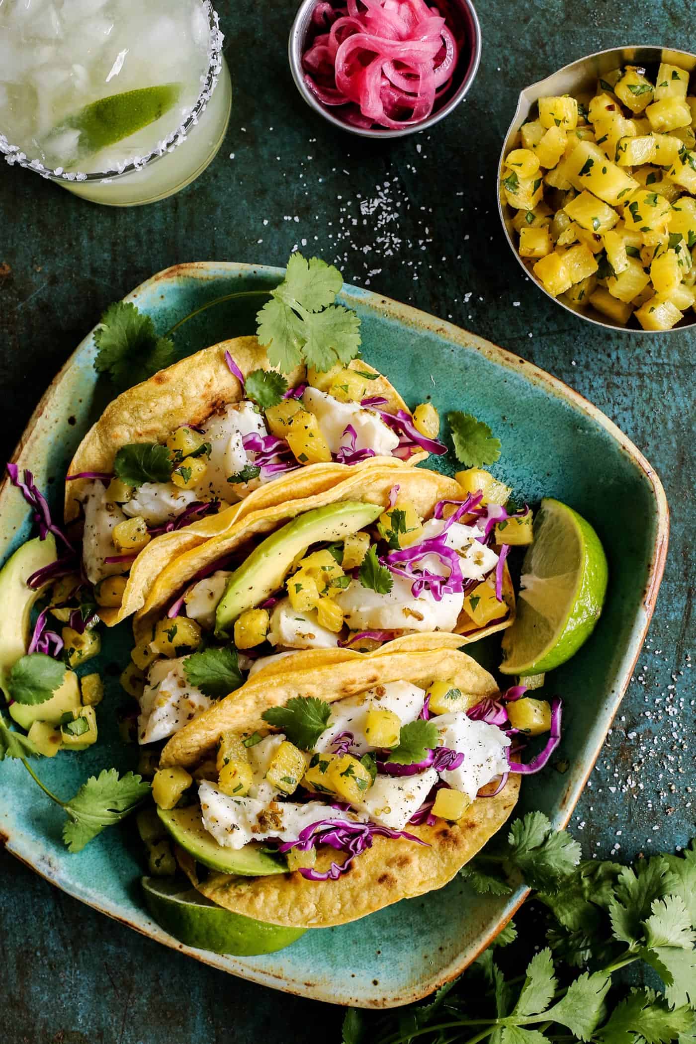 One of the best Fish Tacos Recipes layers caramelized pineapple with lime-spiked white fish, plus creamy avocado and crunchy pickled onions. fish tacos with roasted pineapple on a plate, plus pickled red onions and a margarita