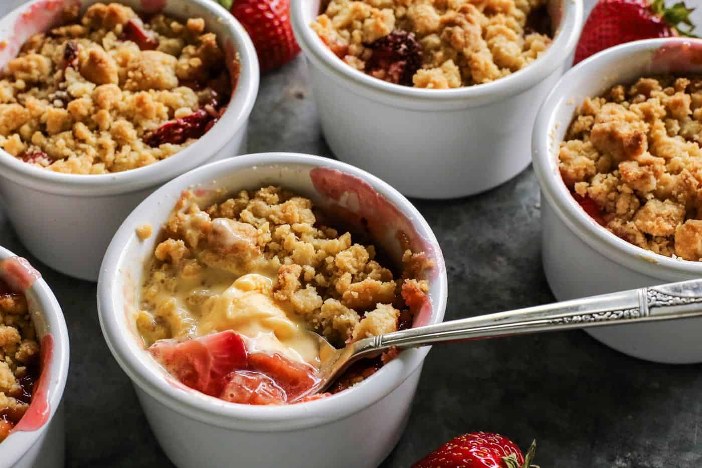 Strawberry Rhubarb Crumble is an easy dessert recipe with a sweet-tart, jammy fruit filling plus a buttery, crispy topping. a spoon dipping into melty vanilla ice cream in a ramekin of rhubarb and strawberry crumble