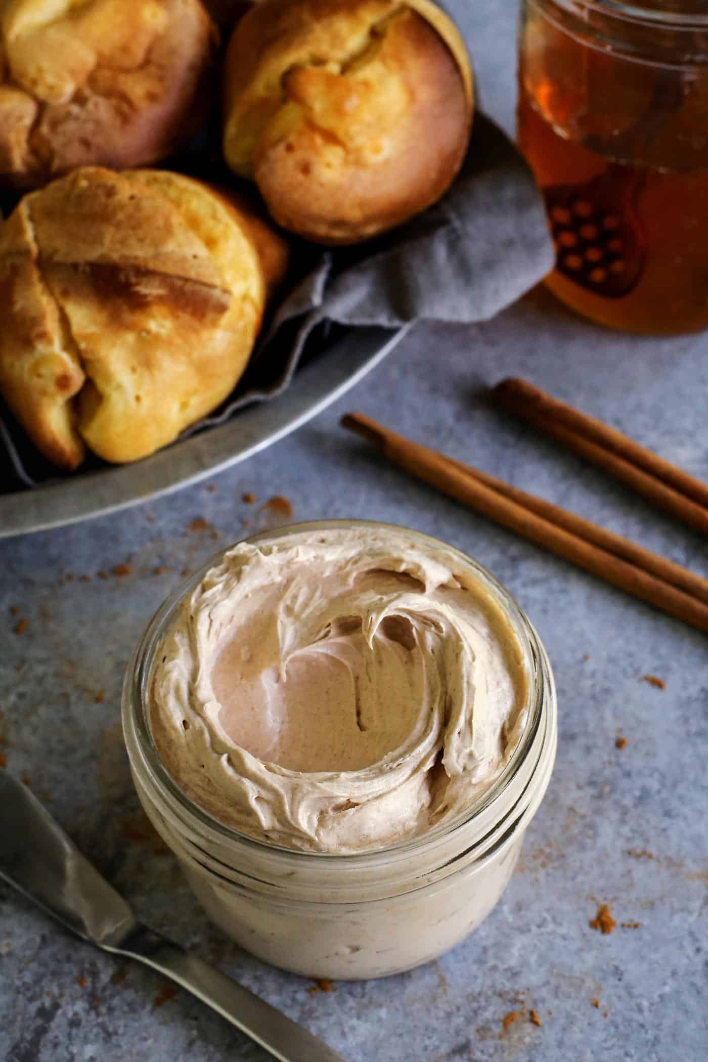 This easy cinnamon honey butter is whipped, fluffy, and even better than Texas Roadhouse butter! Make it with 5 ingredients in 10 minutes! A jar of whipped cinnamon honey butter with a basket of popovers and a jar of honey in the background.