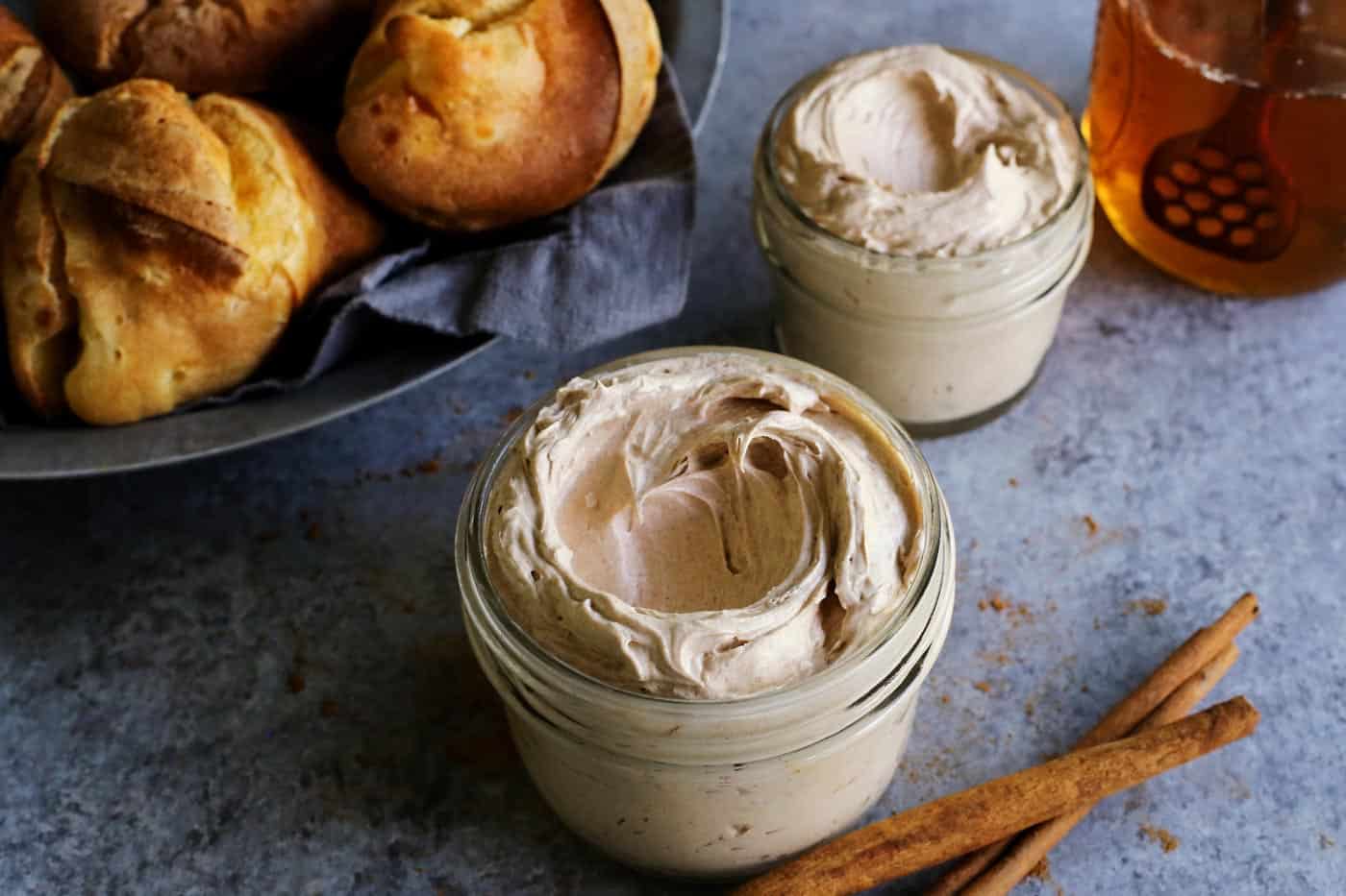 This easy cinnamon honey butter is whipped, fluffy, and even better than Texas Roadhouse butter! Make it with 5 ingredients in 10 minutes! A jar of whipped cinnamon honey butter with a basket of popovers, a second jar of butter, and a jar of honey in the background.