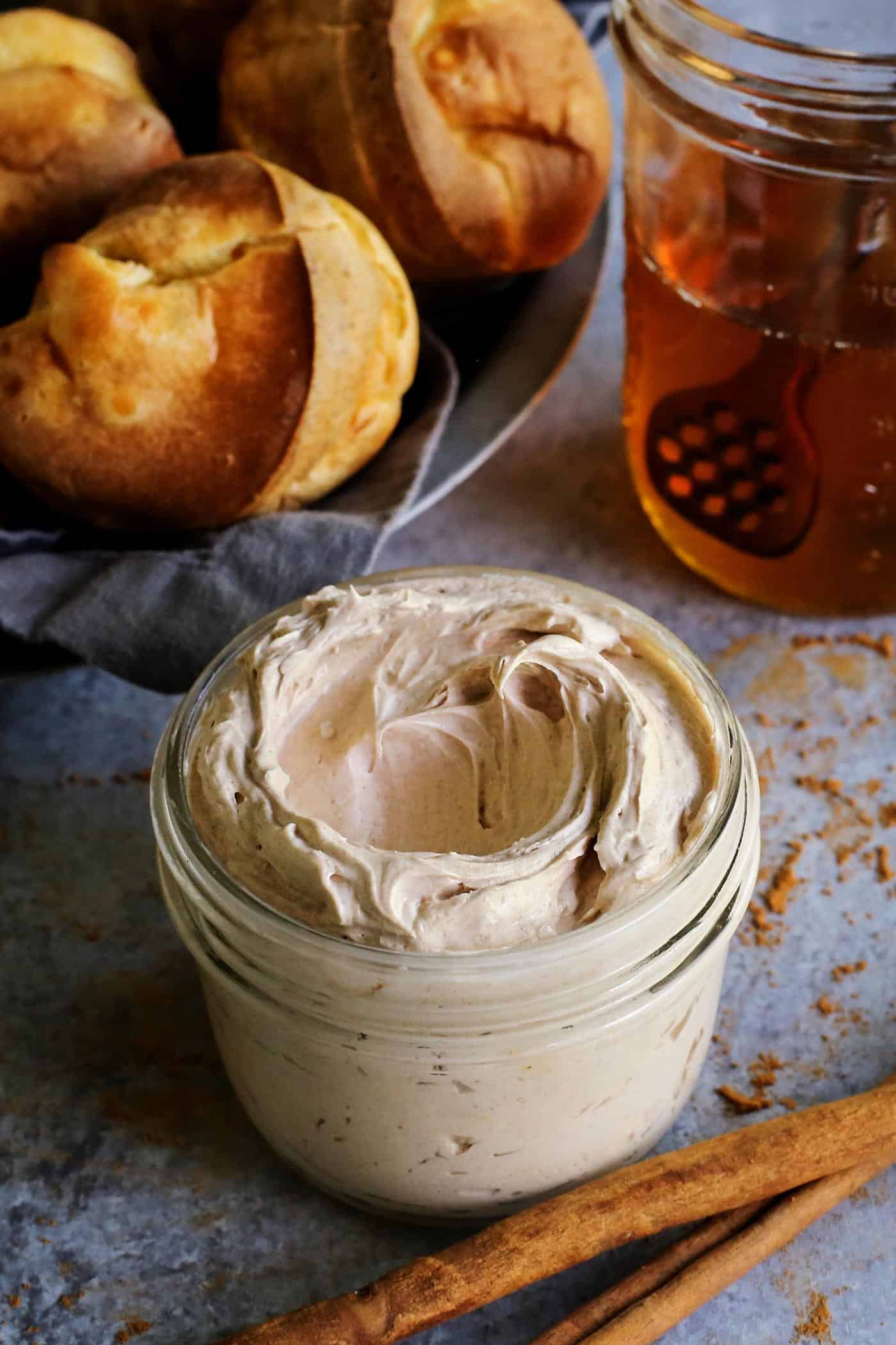This easy cinnamon honey butter is whipped, fluffy, and even better than Texas Roadhouse butter! Make it with 5 ingredients in 10 minutes! A jar of whipped cinnamon honey butter with a basket of popovers and a jar of honey in the background.