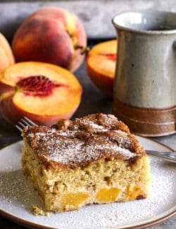 a piece of peach cake on a plate, plus fresh peaches and a cup of coffee