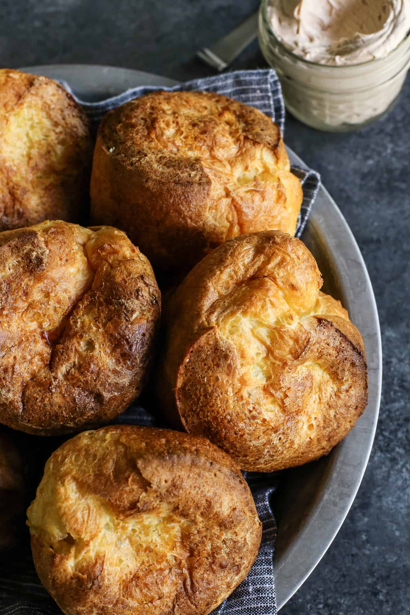 Popovers bake up tall, golden, and airy with crisp edges and soft centers, using only 4 simple ingredients and an easy, foolproof method! Baked popovers in a metal plate, with a jar of cinnamon honey butter on the side.