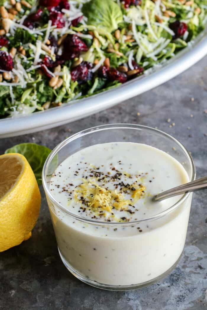 This Shaved Brussels Sprouts Salad has cranberries, sunflower seeds, and Parmesan, with a zippy, creamy dressing. Perfect for the holidays! Lemon yogurt dressing in a glass jar with salad in background.
