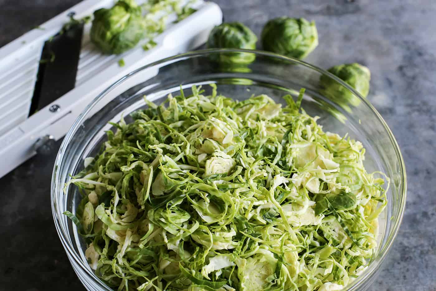 This Shaved Brussels Sprouts Salad has cranberries, sunflower seeds, and Parmesan, with a zippy, creamy dressing. Perfect for the holidays! Shaved Brussels sprouts in a clear bowl, a mandoline cutter in the background.