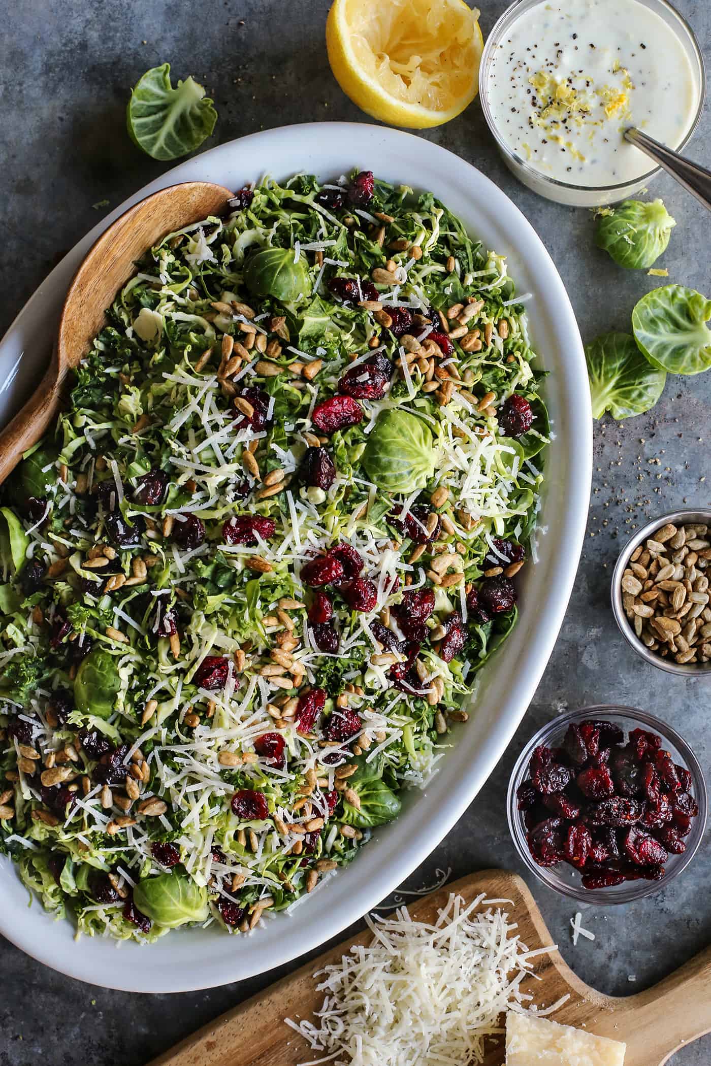 This Shaved Brussels Sprouts Salad has cranberries, sunflower seeds, and Parmesan, with a zippy, creamy dressing. Perfect for the holidays! Shaved Brusssels sprouts salad on an oval white platter with a jar of lemon yogurt dressing.