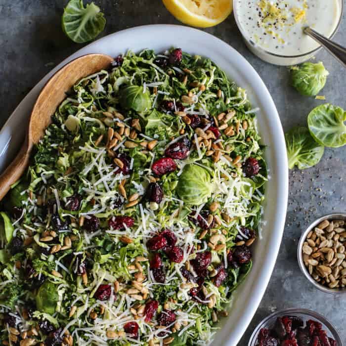 This Shaved Brussels Sprouts Salad has cranberries, sunflower seeds, and Parmesan, with a zippy, creamy dressing. Perfect for the holidays! Shaved Brusssels sprouts salad on an oval white platter with a jar of lemon yogurt dressing.