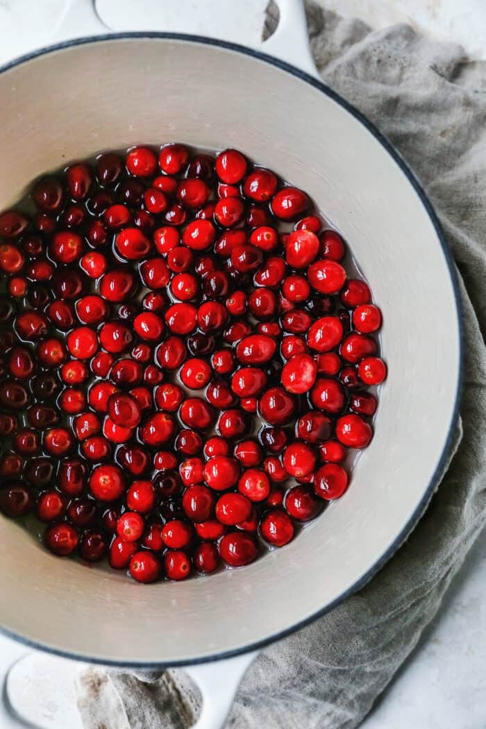 Sugared cranberries are the perfect sparkling finish for all your holiday desserts! All it takes is fresh cranberries, sugar, and water. Overhead view of fresh cranberries soaking in a saucepan of simple syrup.