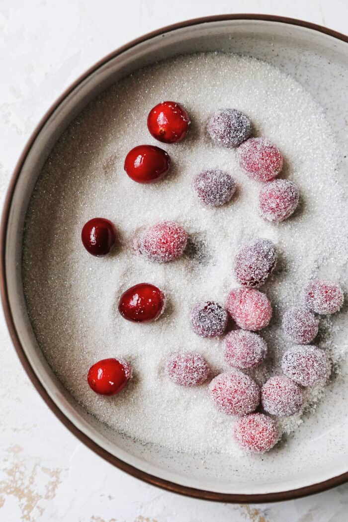 Sugared cranberries are the perfect sparkling finish for all your holiday desserts! All it takes is fresh cranberries, sugar, and water. Close up overhead view of cranberries coated in simple syrup, rolled in sanding sugar in a small bowl.