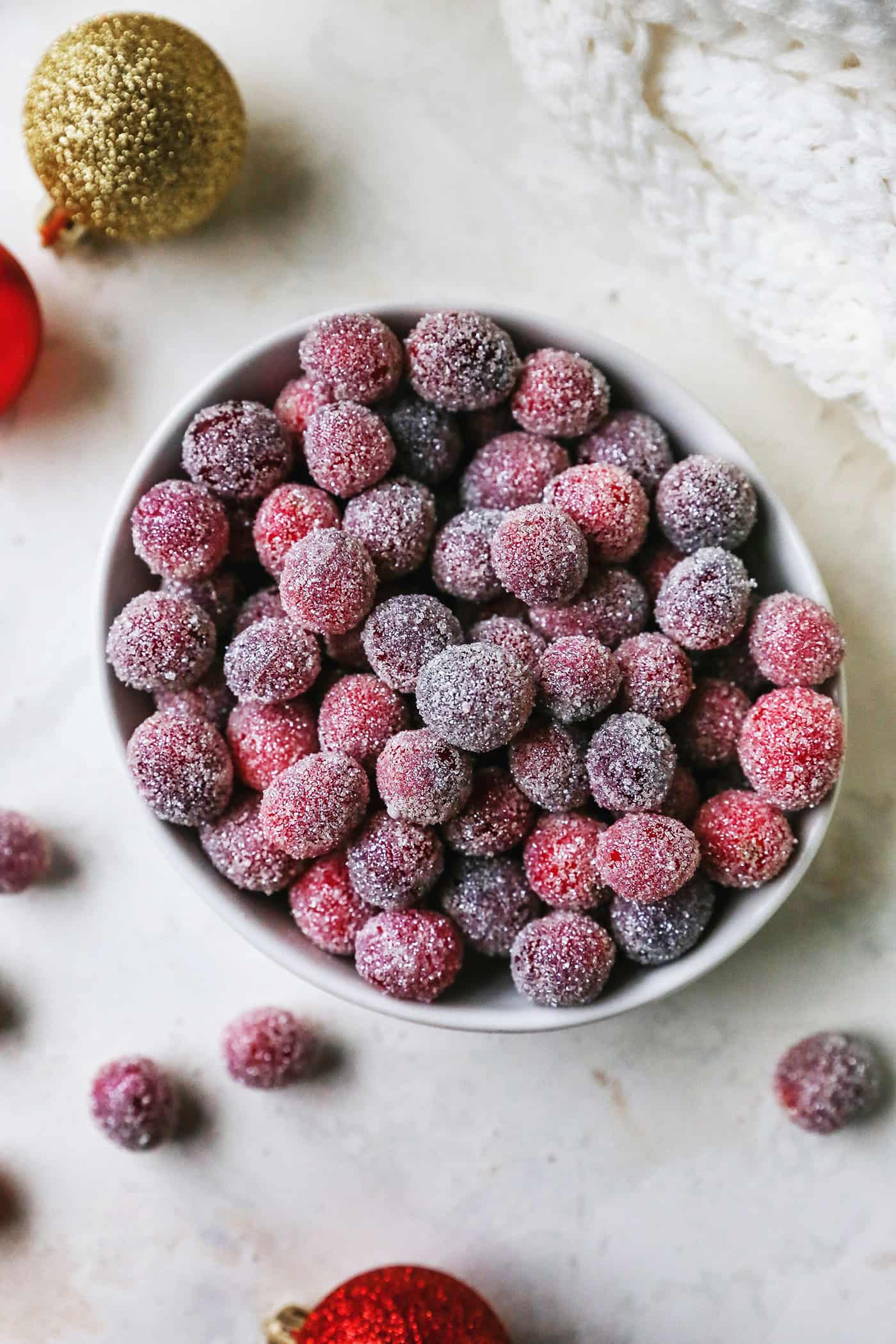 Sugared cranberries are the perfect sparkling finish for all your holiday desserts! All it takes is fresh cranberries, sugar, and water. Overhead view of sugared cranberries in a small bowl next to scattered cranberries and Christmas ornaments.