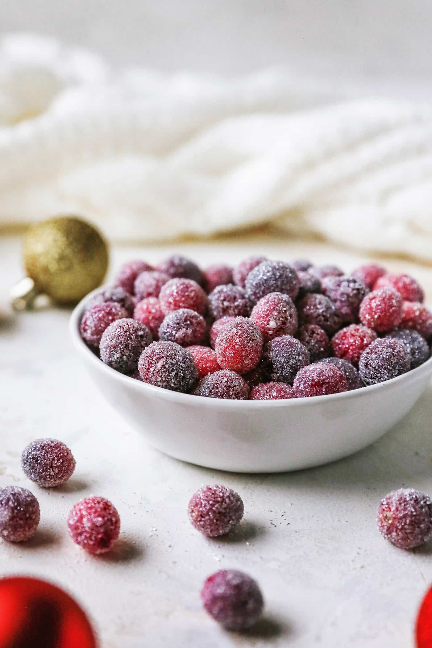 Sugared cranberries are the perfect sparkling finish for all your holiday desserts! All it takes is fresh cranberries, sugar, and water. Sugared cranberries in a small bowl next to scattered cranberries and Christmas ornaments.