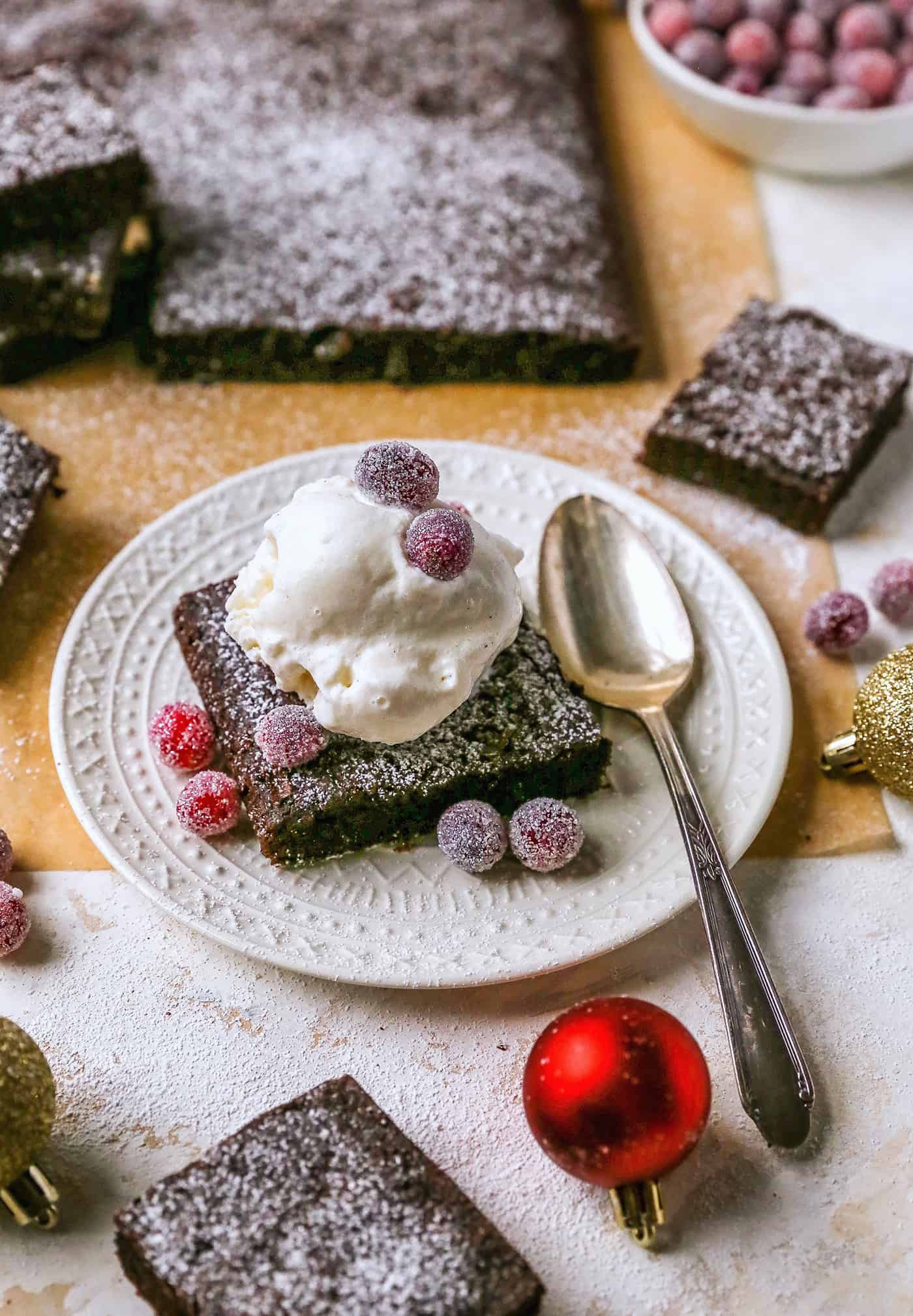 Gingerbread brownies are moist and fudgy, with a touch of molasses and warm spices. A fun holiday twist on a traditional brownie recipe! a gingerbread brownie on a plate, plus Christmas ornaments and sugared cranberries