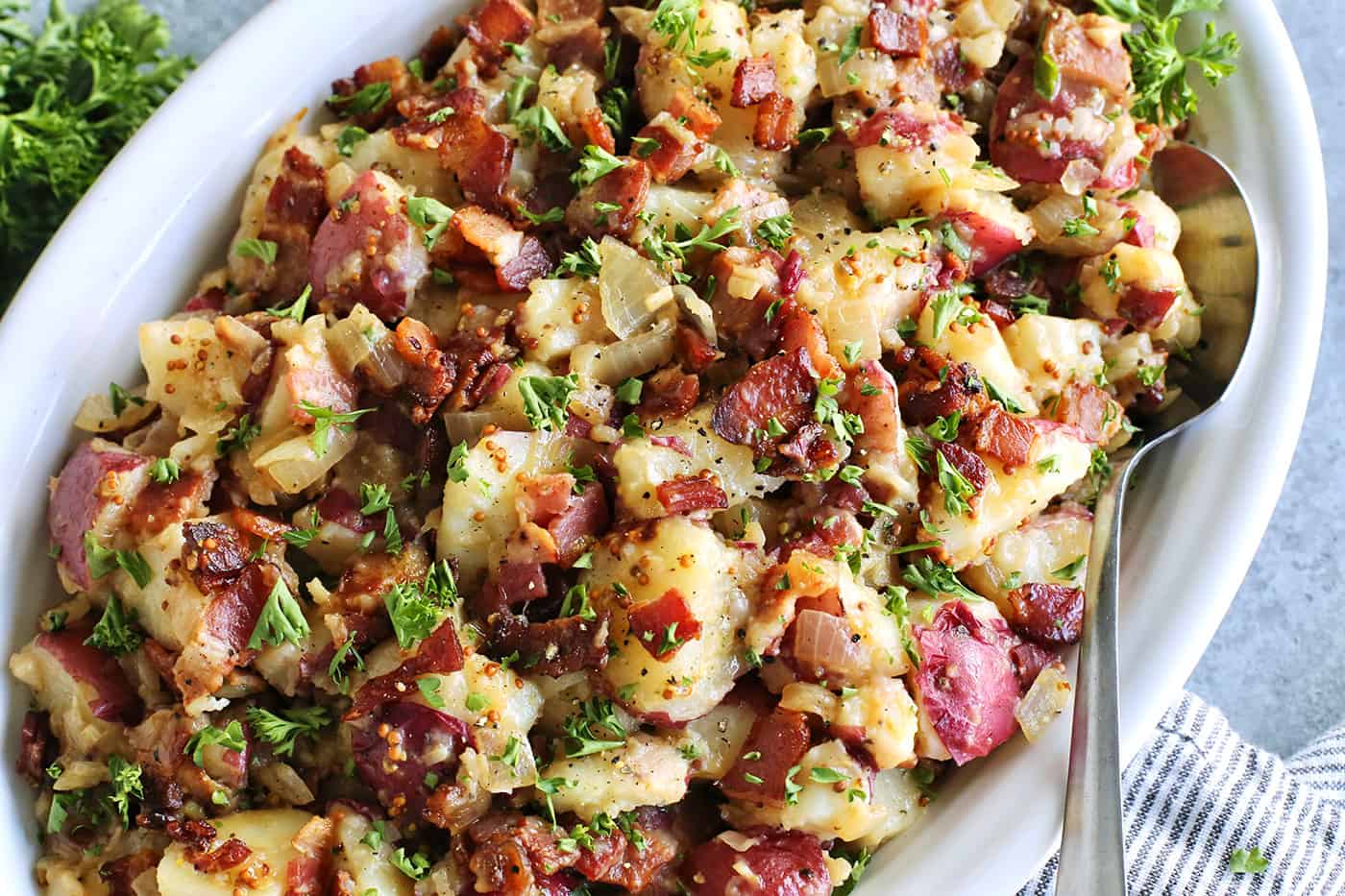 This warm German potato salad recipe combines tender red potatoes with bites of crispy bacon in a tangy vinegar and mustard dressing. a large white platter with potato salad with pieces of bacon
