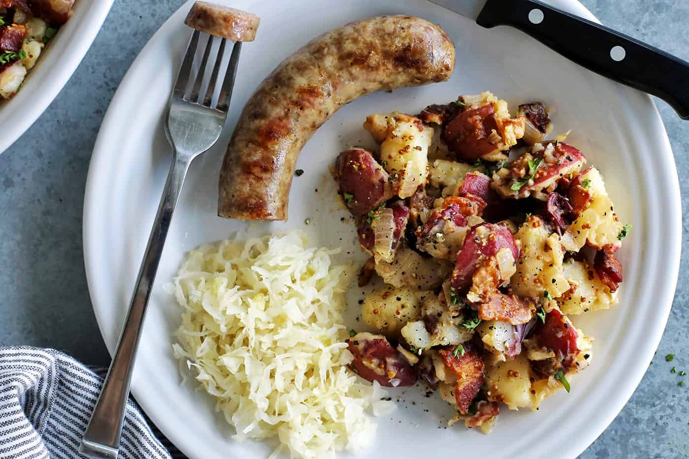 This warm German potato salad recipe combines tender red potatoes with bites of crispy bacon in a tangy vinegar and mustard dressing. a white plate with German potato salad, sauerkraut, and a bratwurst