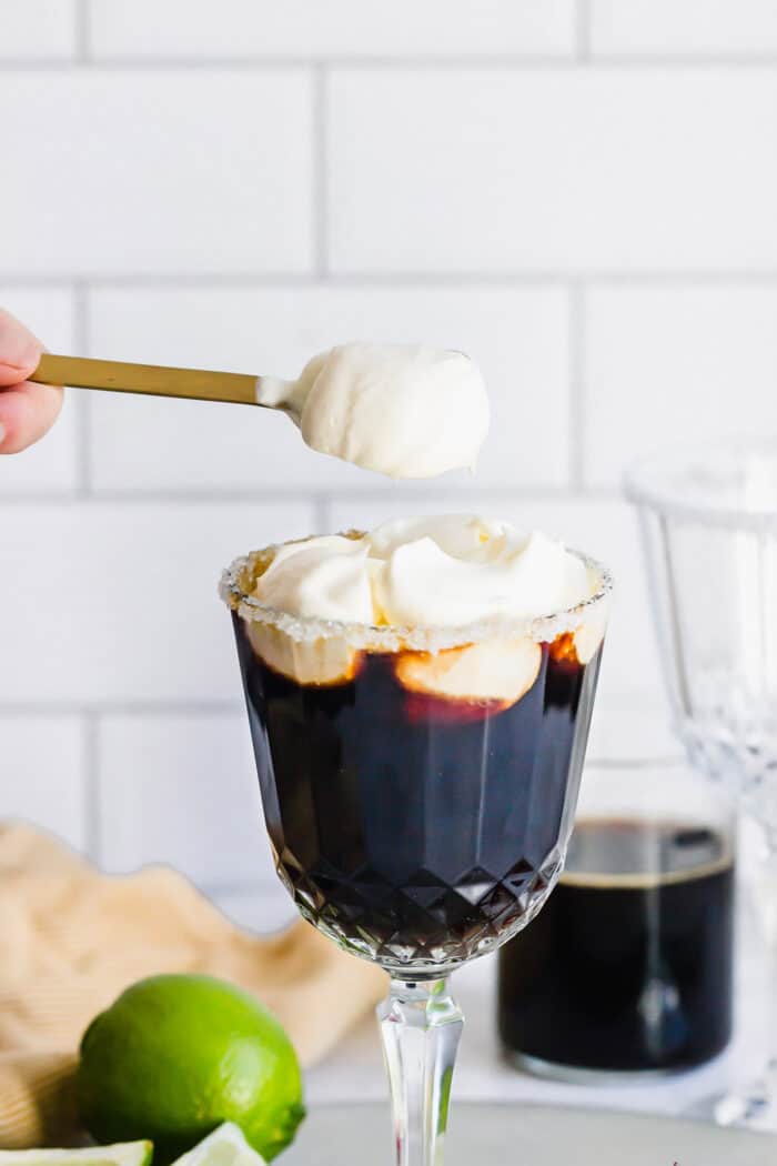 Mexican Coffee is a decadent dessert cocktail that's served hot. With tequila, Kahlua, and whipped cream, this recipe is a fun and beautiful way to top off a meal! adding dollops of whipped cream to a coffee drink in a stemmed glass