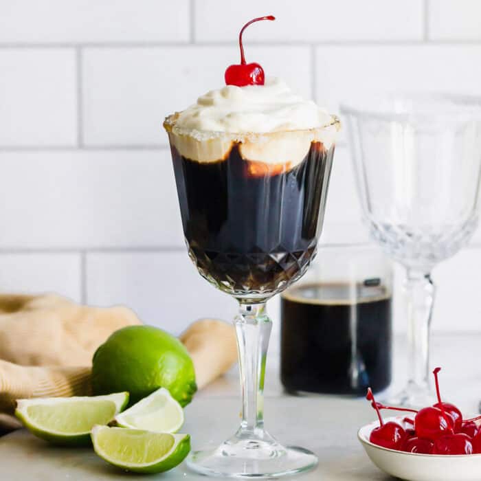 Mexican Coffee is a decadent dessert cocktail that's served hot. With tequila, Kahlua, and whipped cream, this recipe is a fun and beautiful way to top off a meal! Mexican coffee cocktail in a stemmed glass, plus fresh limes and stemmed maraschino cherries