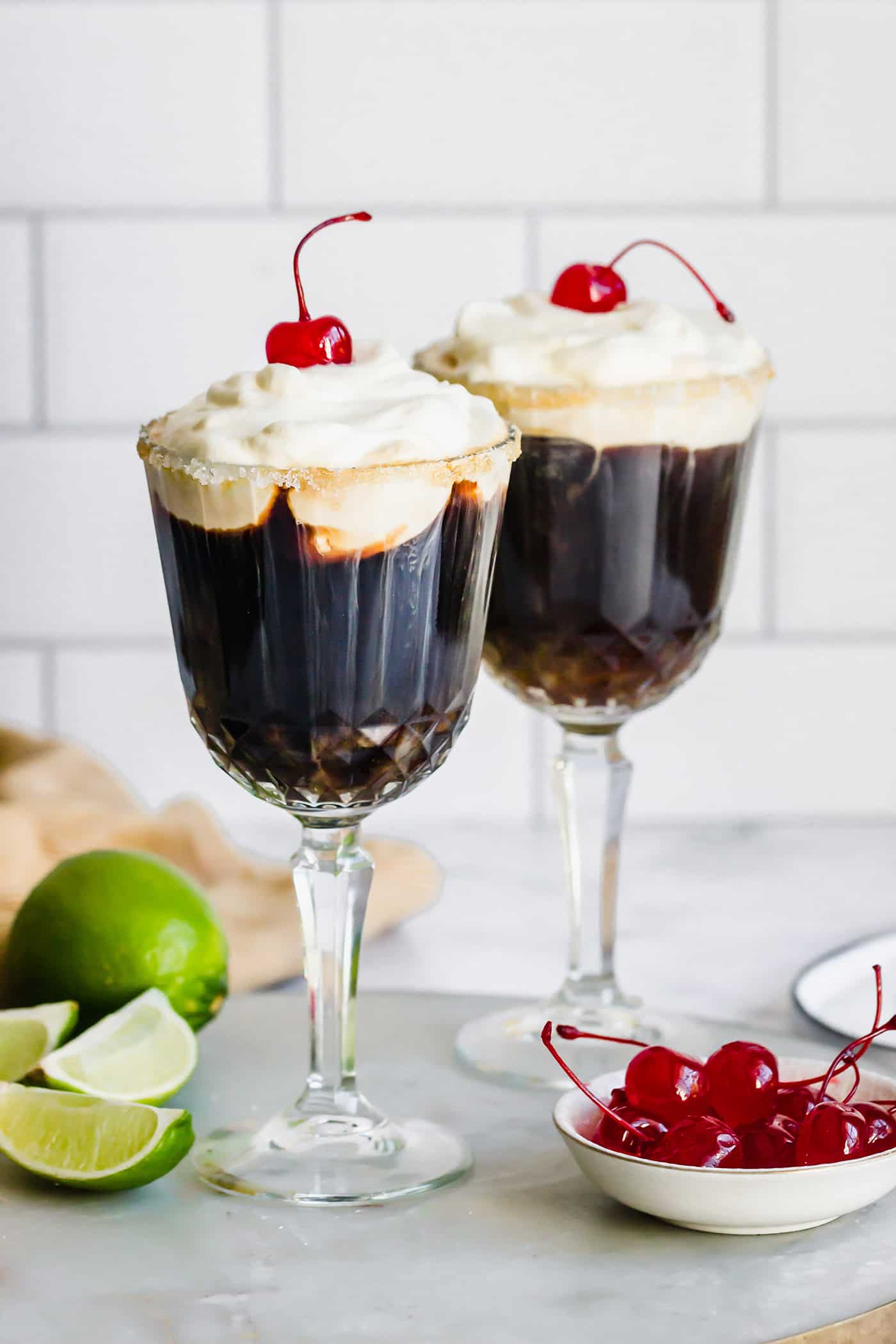 Mexican Coffee is a decadent dessert cocktail that's served hot. With tequila, Kahlua, and whipped cream, this recipe is a fun and beautiful way to top off a meal! two glasses of Mexican coffee in stemmed glasses, with fresh limes and maraschino cherries