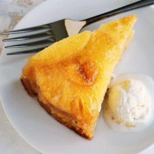 Close up of a slice of homemade pineapple upside down cake next to a scoop of ice cream on a plate, with a fork.