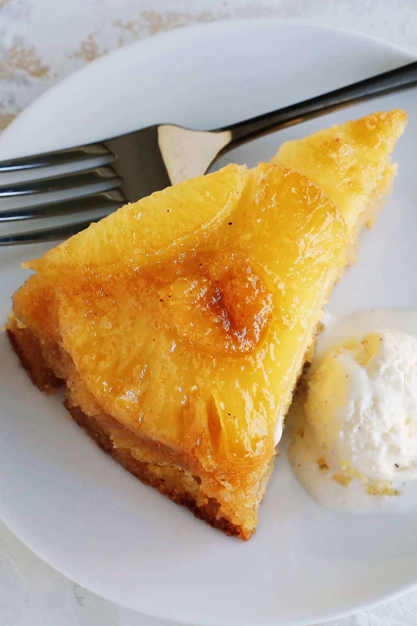 Close up of a slice of homemade pineapple upside down cake next to a scoop of ice cream on a plate, with a fork.