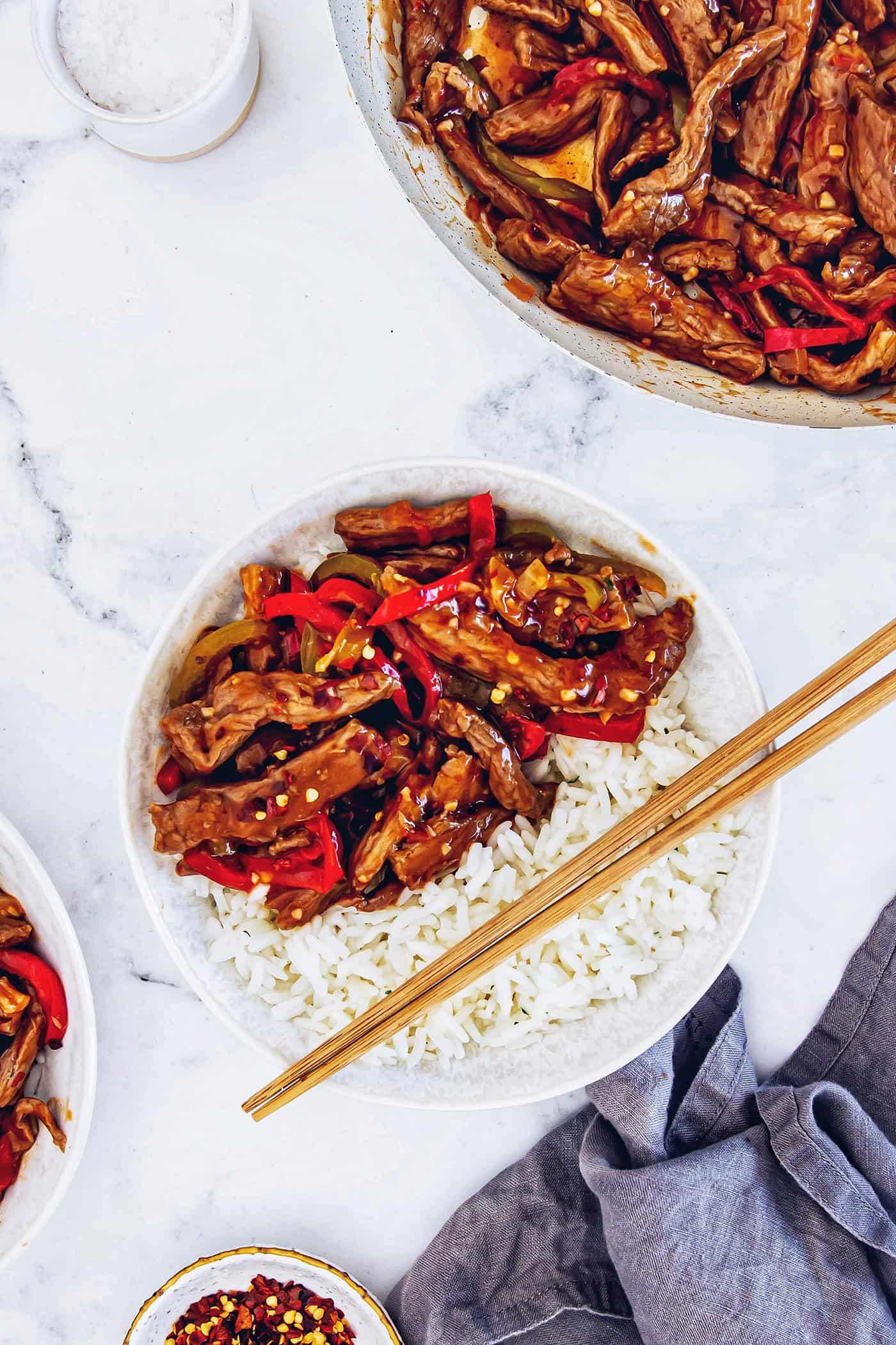 This copycat Beijing beef recipe had tender beef, peppers, and onions tossed in a sweet-spicy homemade sauce. It's even better than takeout! a serving of Beijing beef over white rice in a white bowl, with wood chopsticks