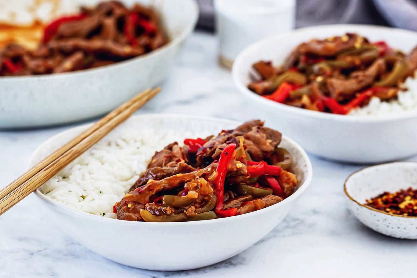 This copycat Beijing beef recipe had tender beef, peppers, and onions tossed in a sweet-spicy homemade sauce. It's even better than takeout! individual bowls of rice, beef, and vegetables with a skillet in the background