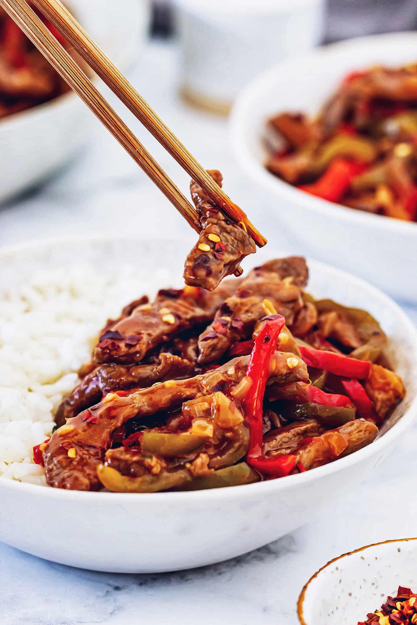 This copycat Beijing beef recipe had tender beef, peppers, and onions tossed in a sweet-spicy homemade sauce. It's even better than takeout! chopsticks holding a piece of Beijing beef over a white bowl