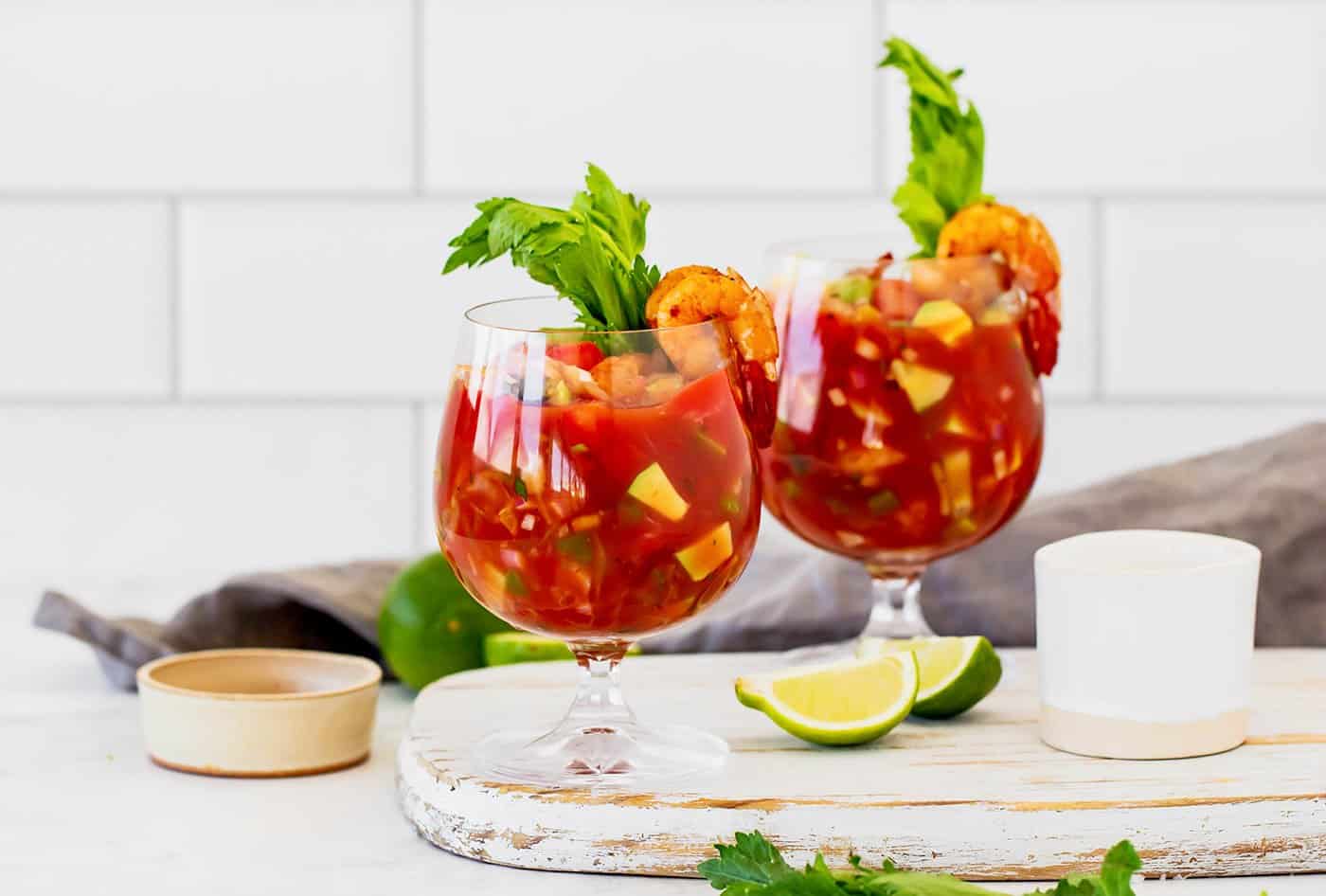 This Mexican shrimp cocktail recipe is a fun twist on a classic appetizer, featuring juicy shrimp and a spicy homemade cocktail sauce! a white cutting board with two stemmed glasses of an appetizer made with tomato juice, lime, and shrimp