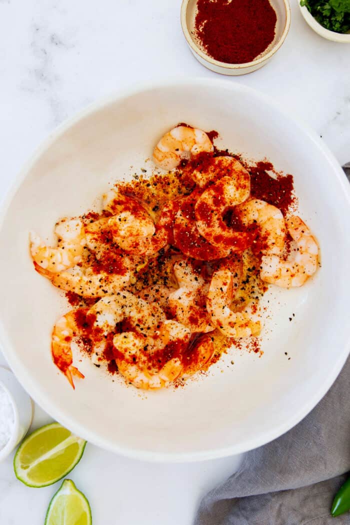 This Mexican shrimp cocktail recipe is a fun twist on a classic appetizer, featuring juicy shrimp and a spicy homemade cocktail sauce! shrimp in a white bowl, sprinkled with chili powder, garlic powder, and smoked paprika