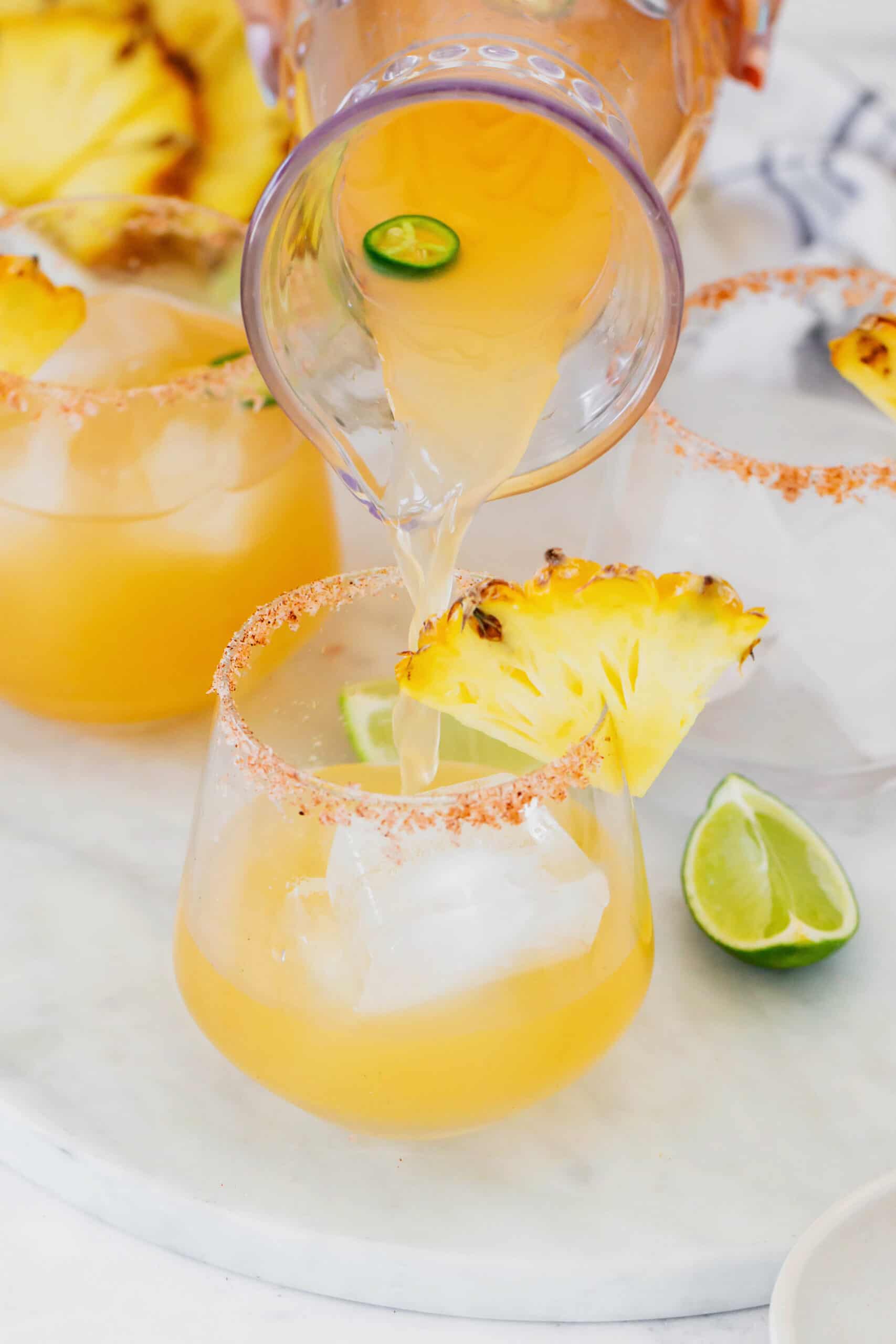 This spicy pineapple jalapeño margarita is a delicious twist on the classic cocktail with blanco tequila, fresh pineapple juice, and jalapeño. pouring pineapple jalapeno margarita mixture from a pitcher to a glass