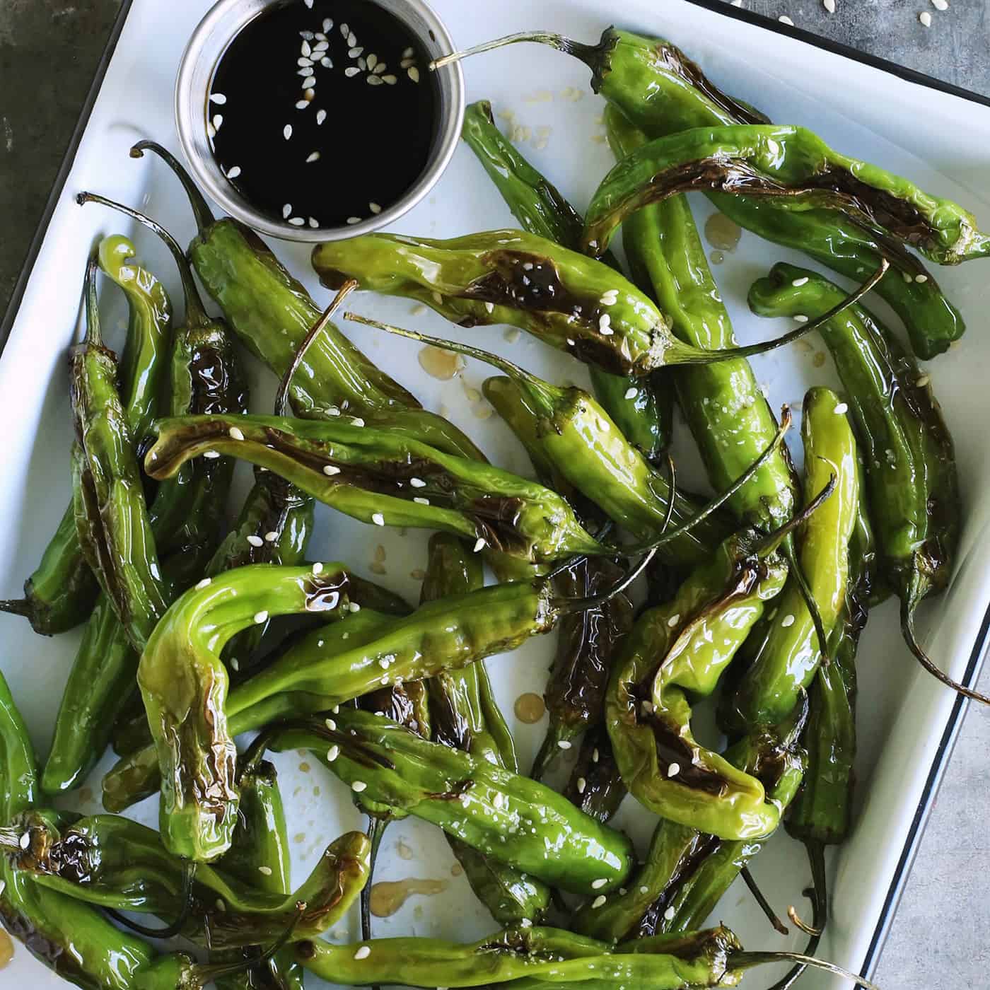 This Blistered Shishito Peppers recipe comes together quickly, with just a few ingredients - one of the easiest appetizers you'll ever make! blistered shishitos on a white enamel tray with soy dipping sauce