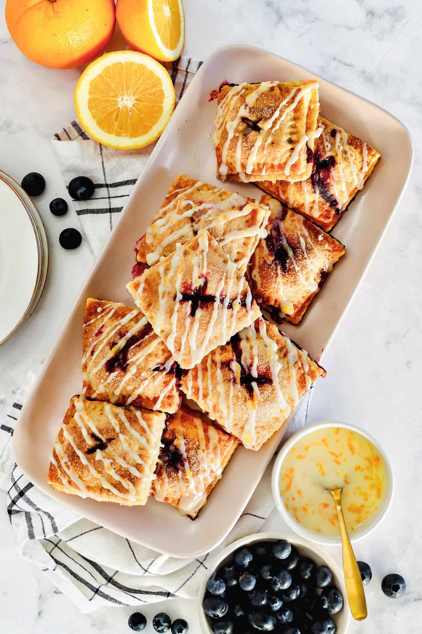 These cute blueberry hand pies combine a flaky pastry crust with sweet blueberry pie filling. It's so much easier than baking a whole pie! Overhead view of a platter of blueberry hand pies drizzled with orange glaze, next to small bowls with pie ingredients.