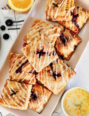 Overhead view of a platter of blueberry hand pies drizzled with orange glaze, next to small bowls with pie ingredients.