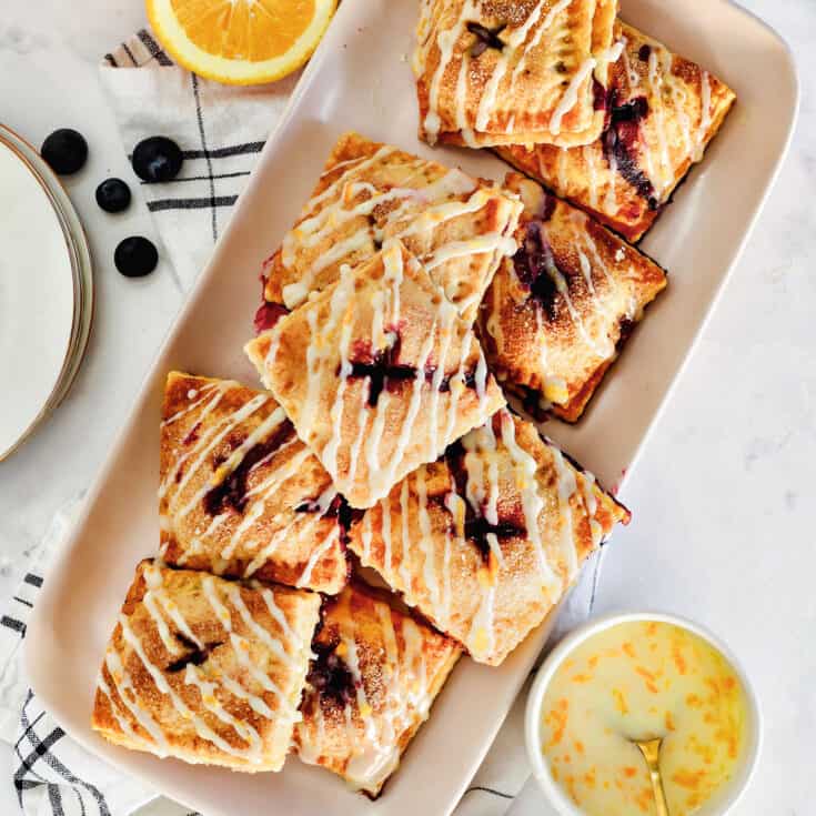 Overhead view of a platter of blueberry hand pies drizzled with orange glaze, next to small bowls with pie ingredients.