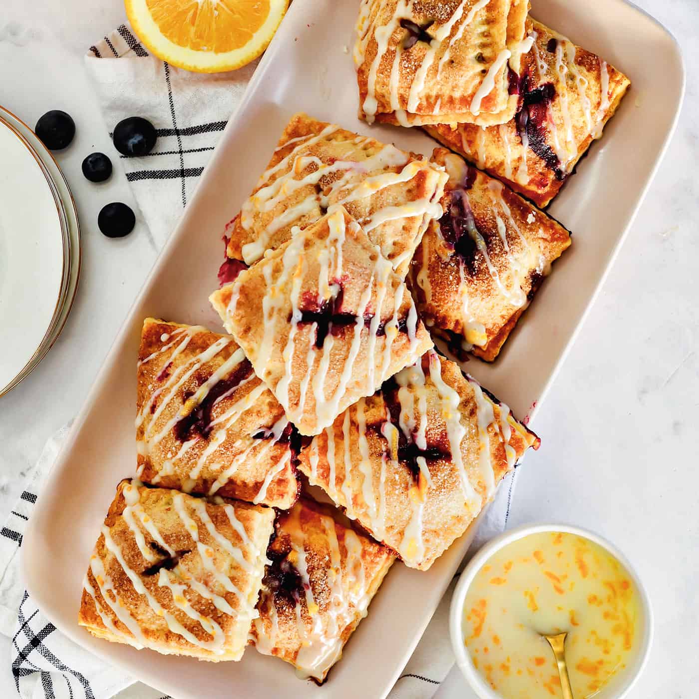 These cute blueberry hand pies combine a flaky pastry crust with sweet blueberry pie filling. It's so much easier than baking a whole pie! Overhead view of a platter of blueberry hand pies drizzled with orange glaze, next to small bowls with pie ingredients.