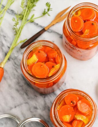 three jars of pickled carrots and a couple whole carrots with their green tops