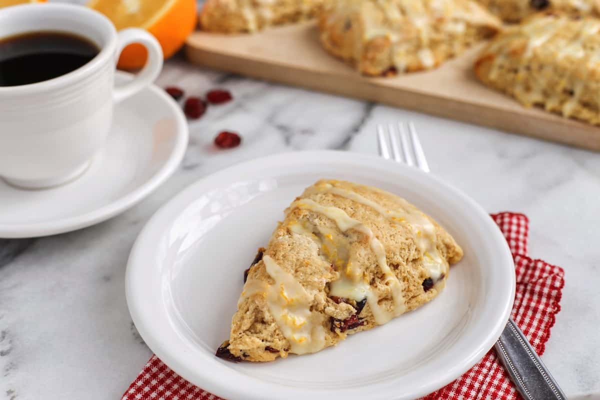 These homemade cranberry orange scones are perfectly flaky and great for a weekend breakfast or holiday brunch. They're easy to make too! A cranberry orange scone drizzled with glaze on a white plate, with more scones on a wooden cutting board next to a cup of coffee in the background.
