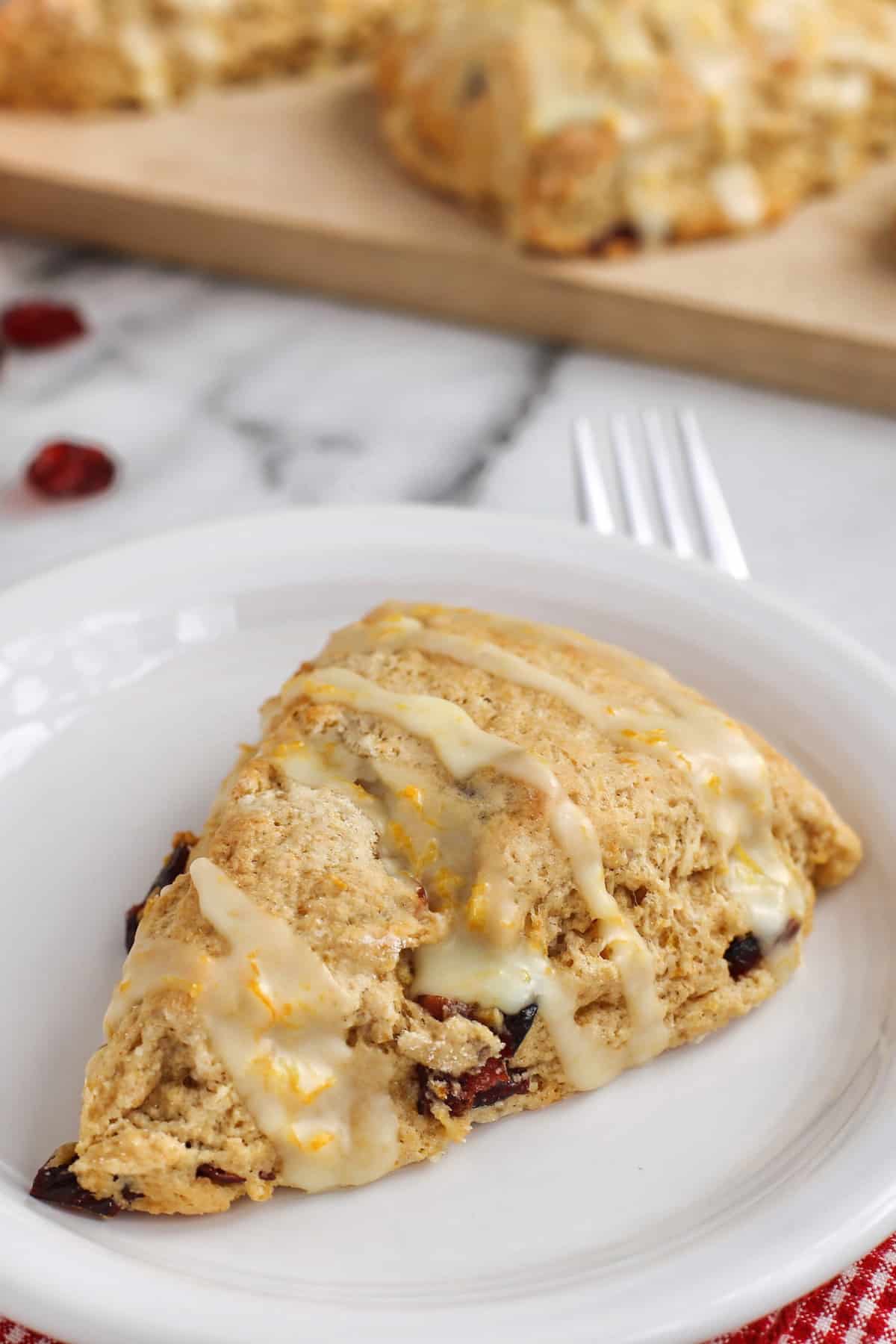 These homemade cranberry orange scones are perfectly flaky and great for a weekend breakfast or holiday brunch. They're easy to make too! A cranberry orange scone drizzled with glaze on a white plate, with more scones on a wooden cutting board in the background.