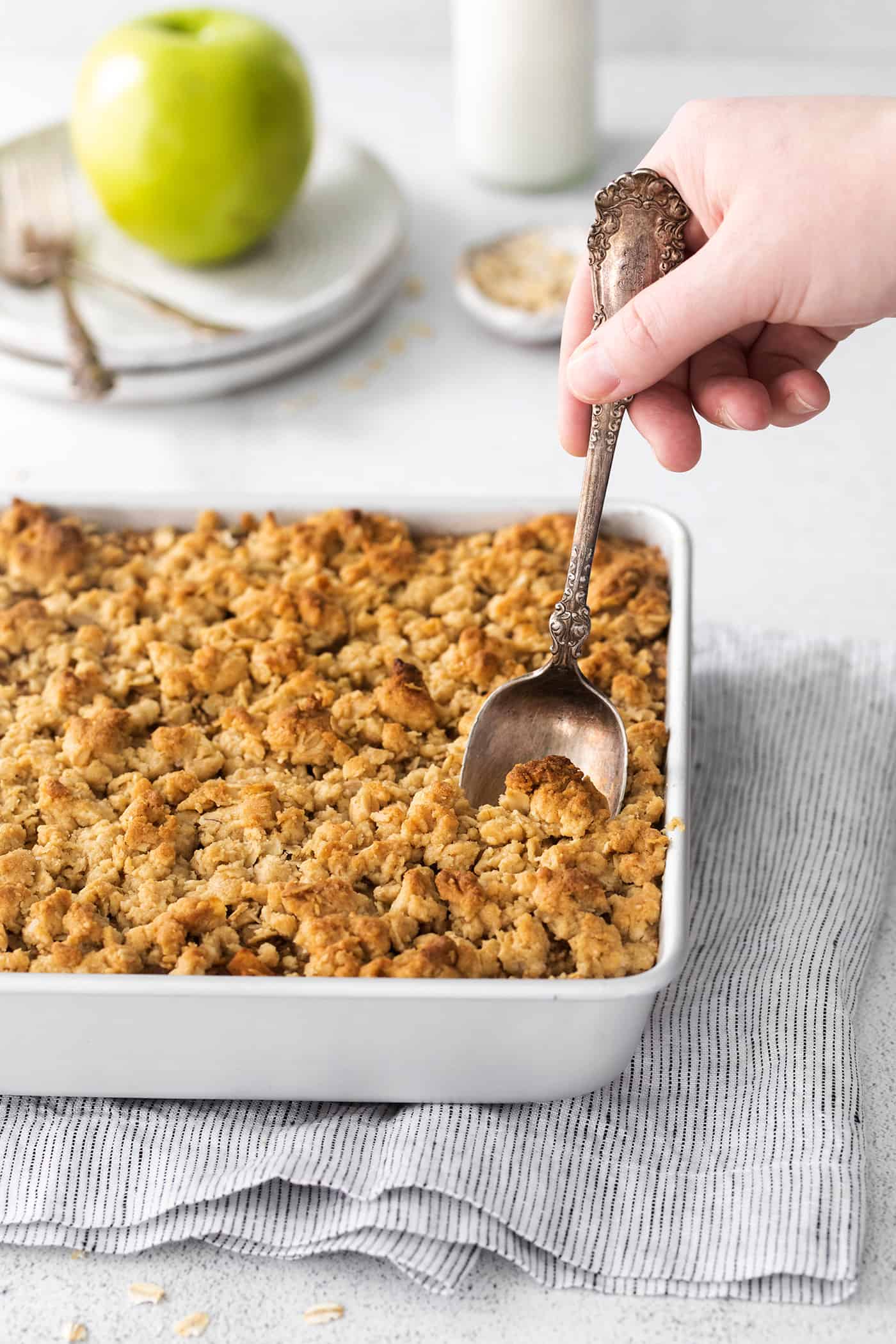 Overnight baked apple oatmeal is an irresistible make-ahead breakfast, topped with crunchy streusel and studded with sweet cinnamon apples. A hand sticking a spoon into overnight baked apple oatmeal in a ceramic baking dish on a kitchen counter.