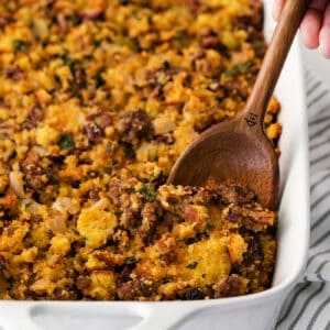 A wooden spoon serving cornbread sausage dressing from a white casserole dish.