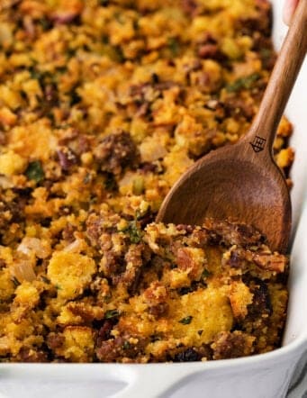 A wooden spoon serving cornbread sausage dressing from a white casserole dish.