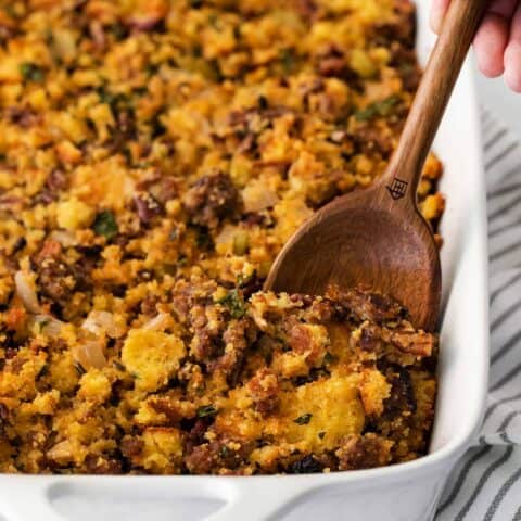 A wooden spoon serving cornbread sausage dressing from a white casserole dish.