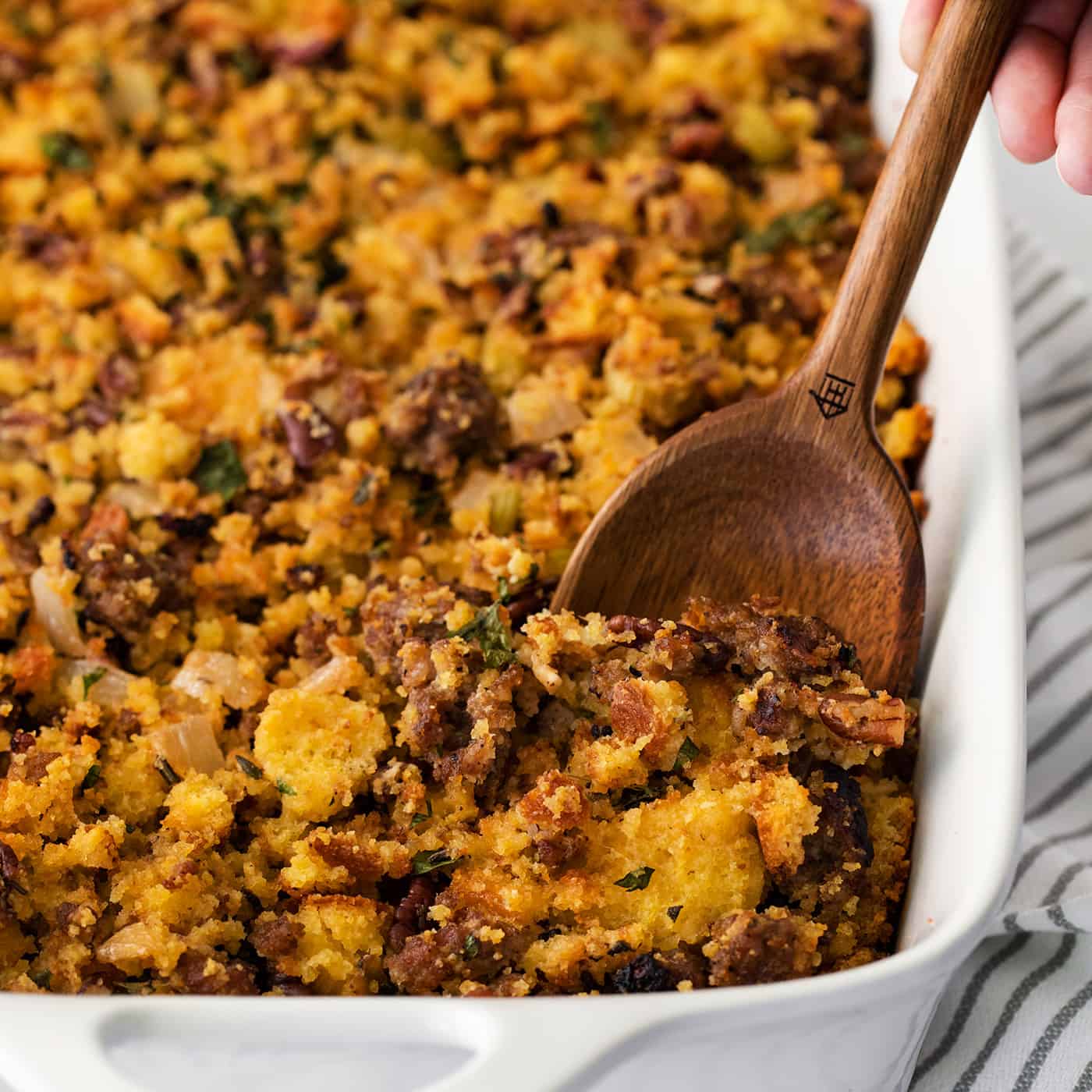 A wooden spoon serving cornbread sausage dressing from a white casserole dish.
